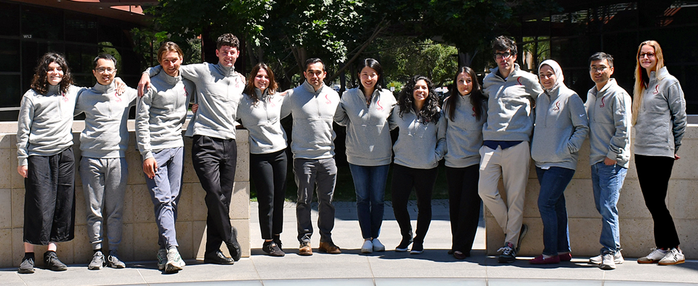 13 postdocs in matching Bio-X logo sweaters standing in the Courtyard with their arms around each other.