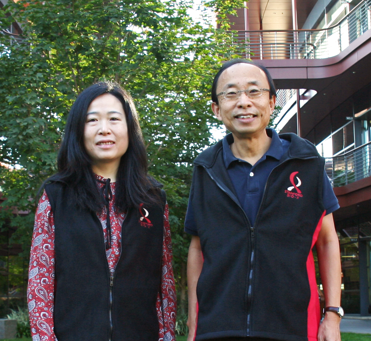 Photo of female Asian faculty member and male Asian faculty member standing side by side at the Clark Center, wearing matching vests with the Bio-X logo.