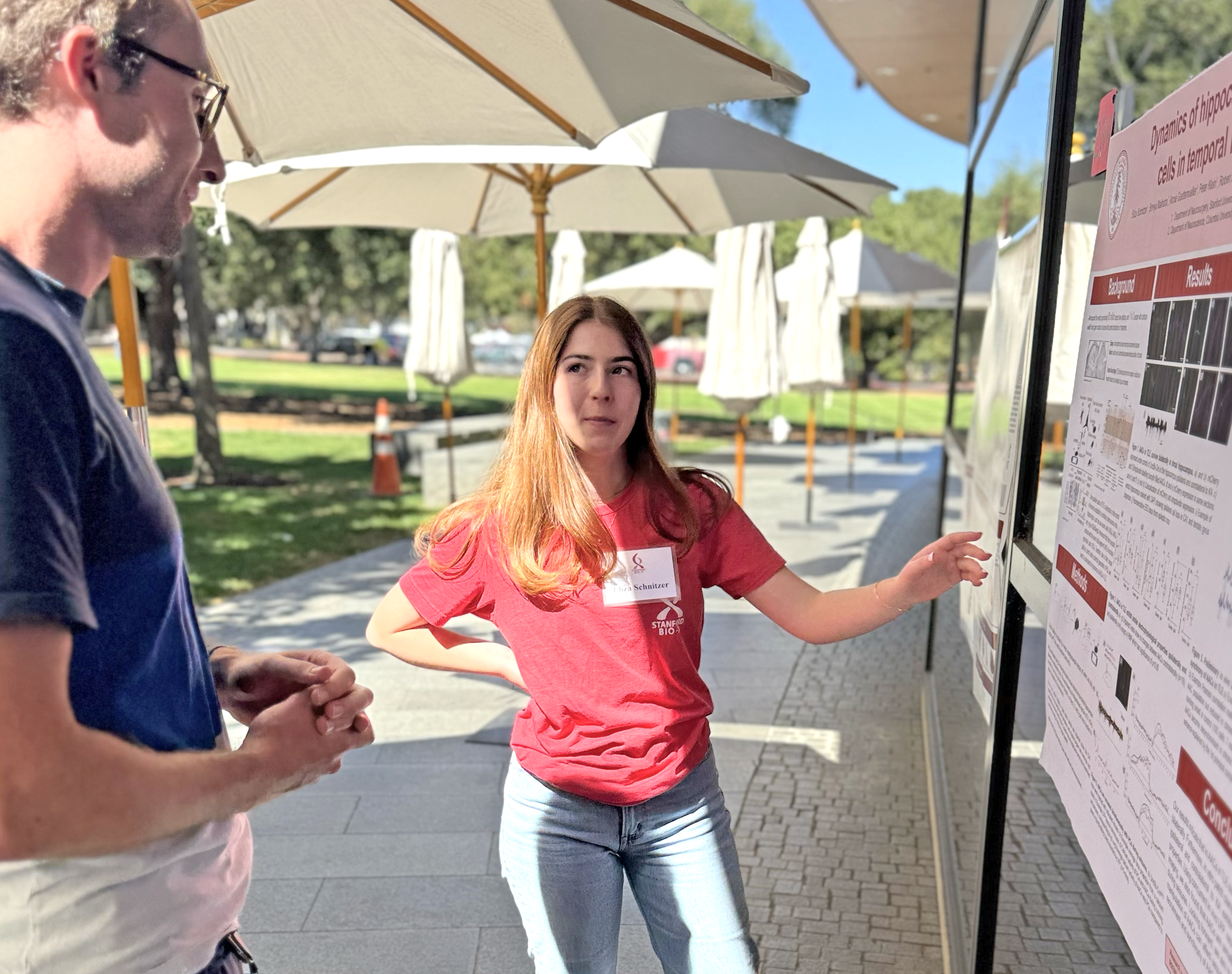 Outdoor photo of a male Asian undergraduate in a red Bio-X T-shirt showing a poster to another researcher.