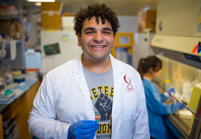 Indoor photo of a Black male undergraduate student in the lab, wearing a Bio-X lab coat and smiling at the camera.