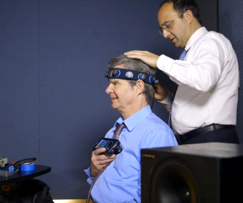Indoor photo of two men, one putting a complex headband on the other, who holds up a readout device.