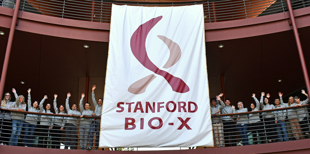 Postdocs and staff wearing matching jackets waving down, with a large Bio-X logo sign in the center.