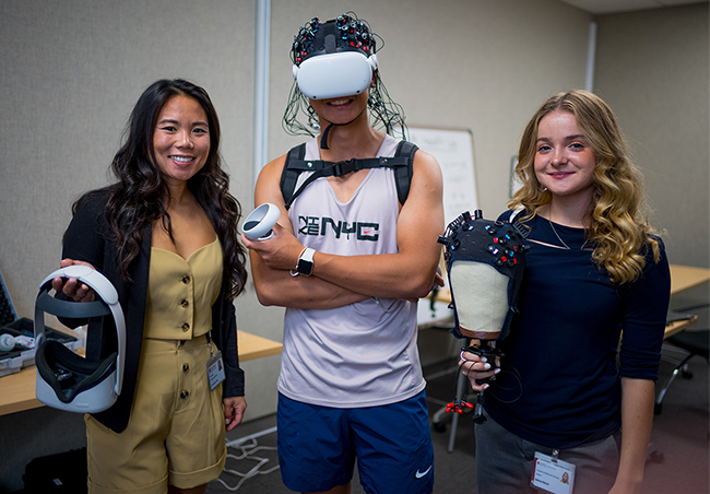 Indoor photo of an Asian female postdoctoral scholar, an Asian male undergraduate, and a Causasian female undergraduate all holding or wearing various brain scanning helmets and devices.