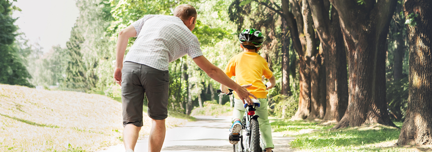 Photo of a father helping his son to learn to ride a bike in a wooded area.