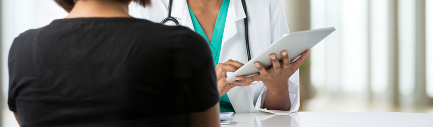 Photo over a female patient's shoulder of a female doctor holding up a clipboard as they talk across a table.