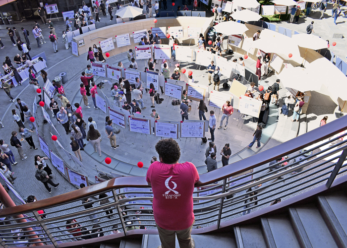 Outdoor photo of a student leaning over a staircase railing, watching people browse scientific posters in the Clark Courtyard.