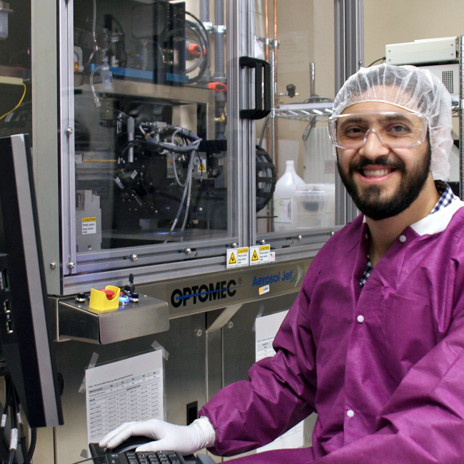 Photo of graduate student Abdul Obaid in the lab, dressed in clean room attire and seated beside a large piece of equipment.