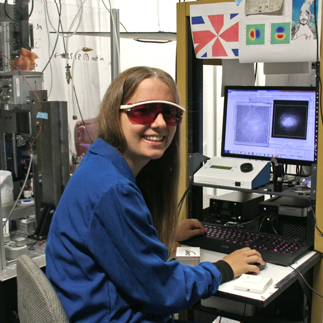 Photo of graduate student Annina Sartor in the lab, wearing red-lensed safety goggles, sitting at a computer in front of a laser apparatus.