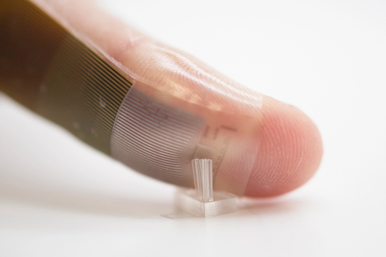 Photo of a person's fingertip with electrode patches stuck on, conforming to the finger. The fingertip is pressing against a translucent object with upward protruding stems.