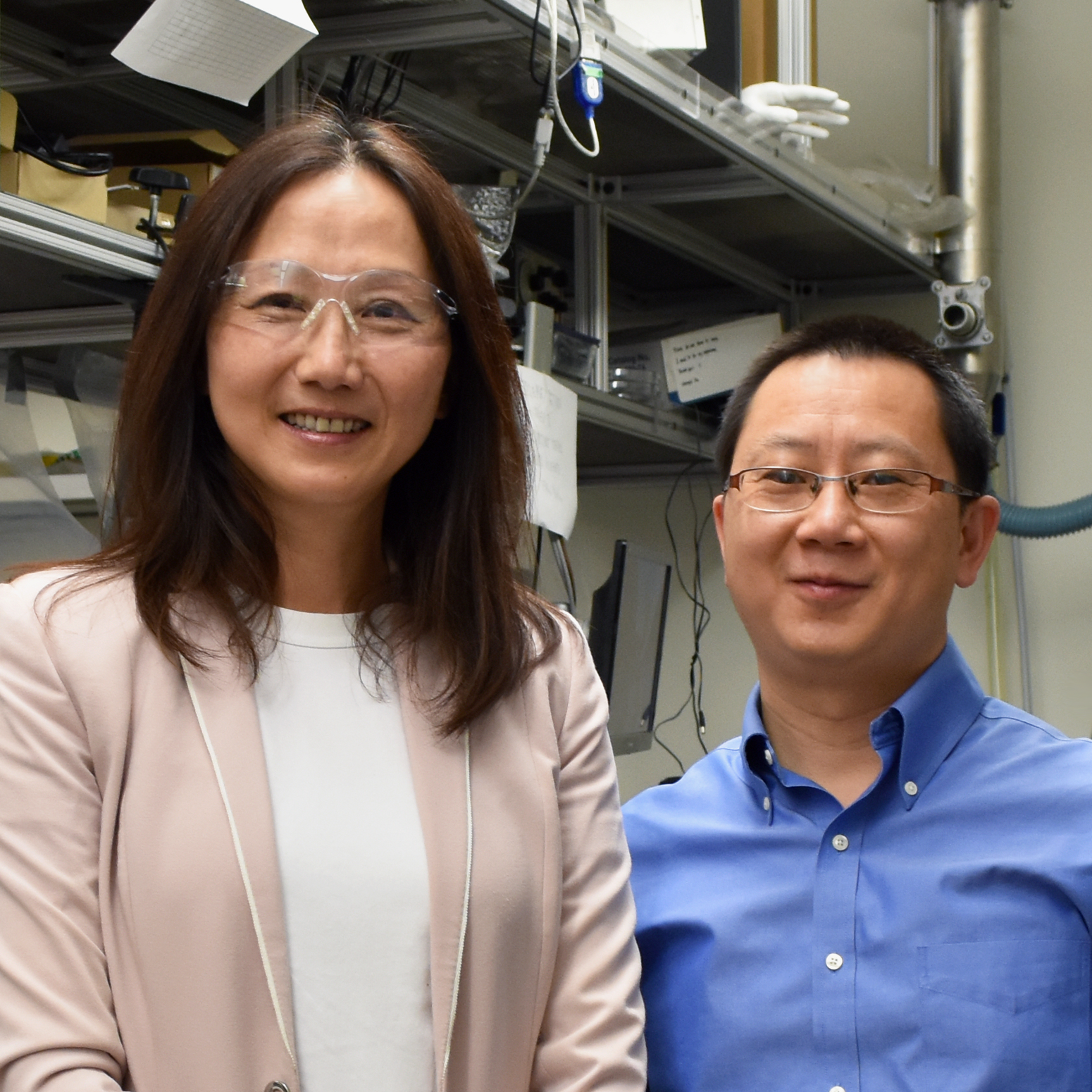 Indoor photo of an Asian female faculty member with long black hair standing on the left, wearing safety goggles and a pale pink blazer and a white shirt, and a smiling Asian male faculty member with short black hair and glasses wearing a blue button up shirt, standing inside of an engineering laboratory with shelves and equipment in the background..