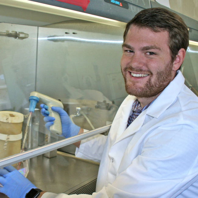 Photo of graduate student Bauer LeSavage in the lab, pipetting inside a tissue culture hood.