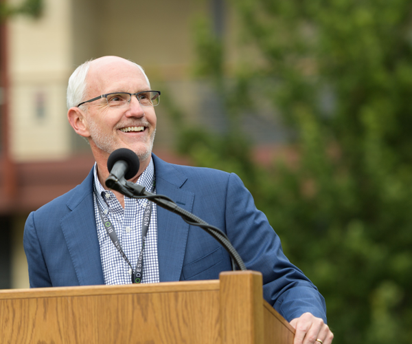 Outdoor photo of a white male professor standing at a lectern in front of trees and smiling.