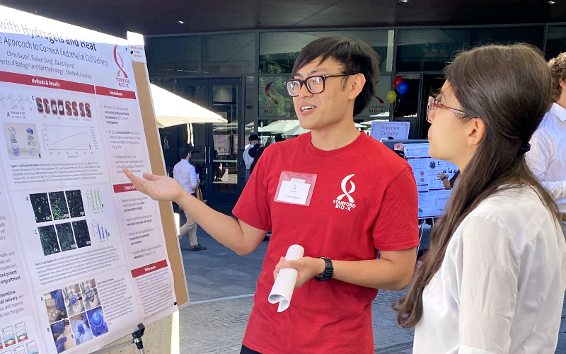 Outdoor photo of a male Asian undergraduate in a red Bio-X T-shirt showing a poster to another researcher.