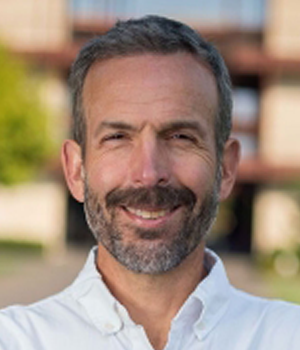 Outdoor headshot photo of smiling male faculty member with short hair and a beard.