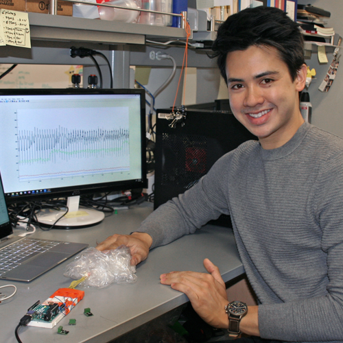 Photo of undergraduate student Cody Carlton sitting at a desk, holding a small device with numerous attachments, which is showing a heartbeat EKG-like reading on a computer monitor behind him.