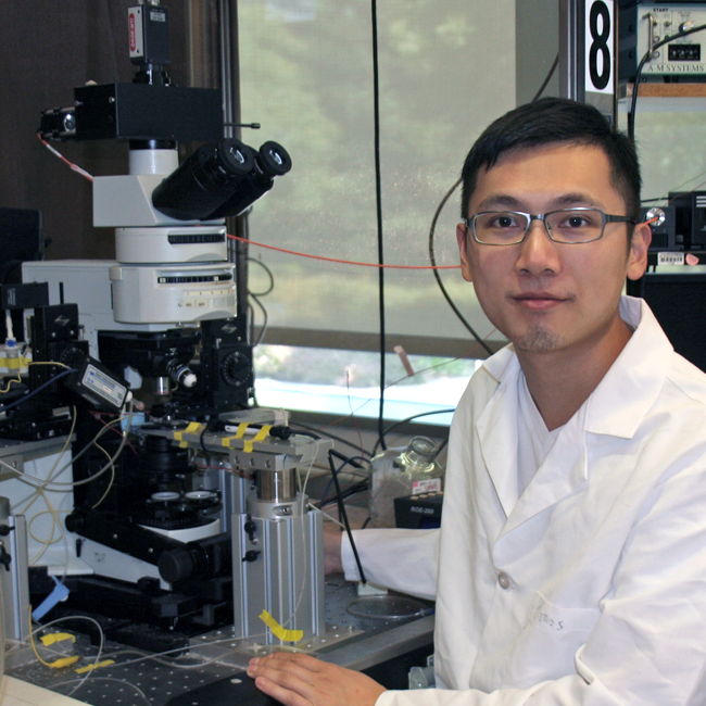 Photo of graduate student Cosmos Wang in the lab, seated next to a large microscope.