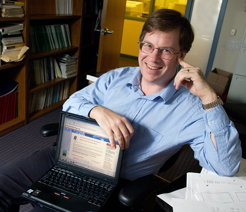 Photo of Dr. David Dill in his office, with a laptop computer in his lap and notes on the desk nearby.