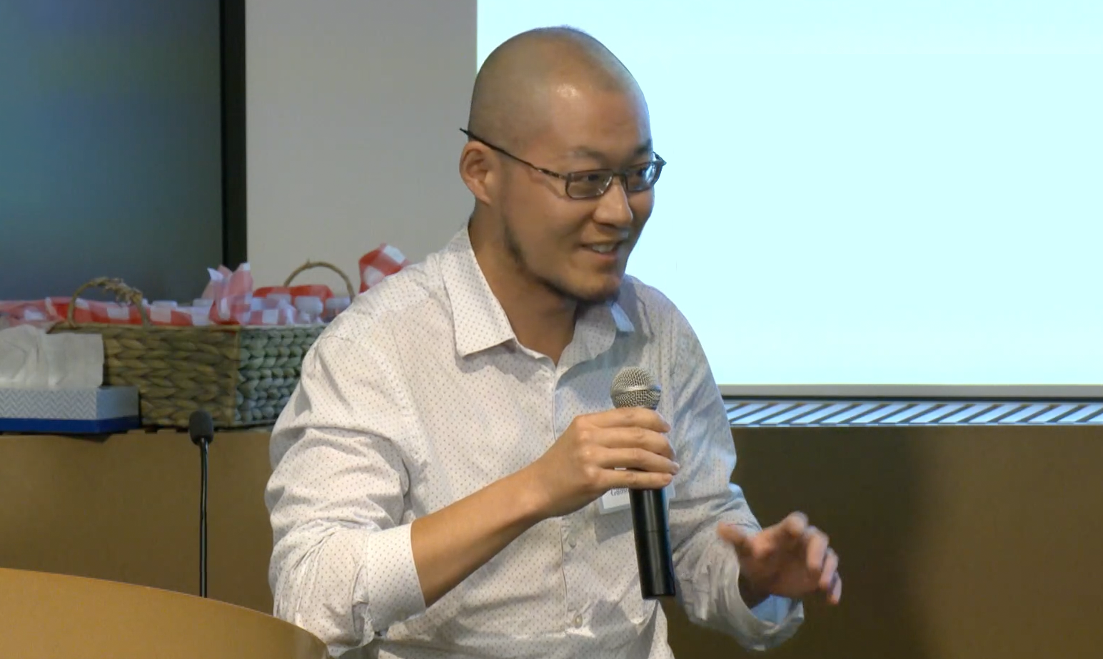 Indoor photo of male Asian faculty member holding a microphone in front of a projector screen.
