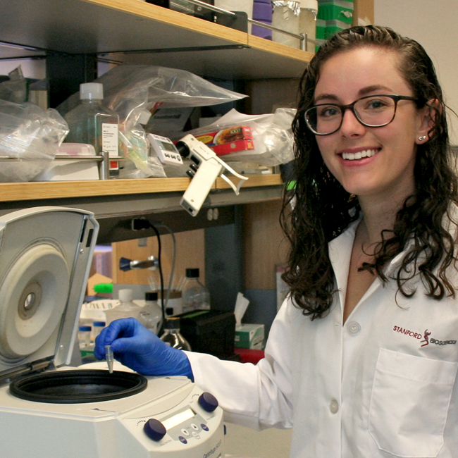 Photo of graduate student Nina Horowitz in a wet lab, wearing glasses and a lab coat and gloves, putting a sample into a centrifuge.