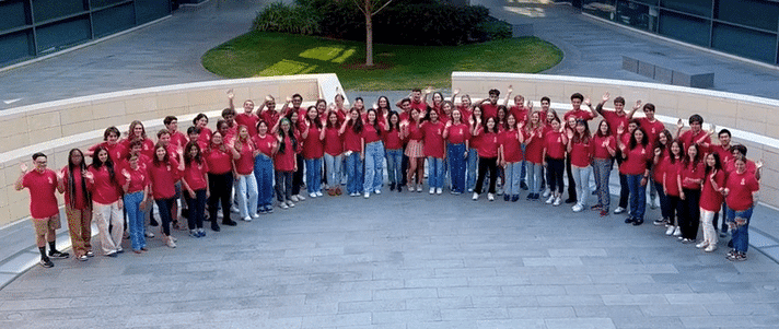 Animated gif of 70 undergraduate students outdoors, wearing matching light red T-shirts, waving up at the camera.