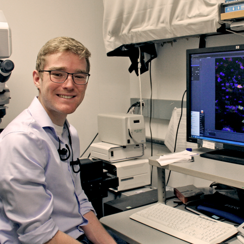 Photo of undergraduate student Jacob Greene sitting in lab space, with microscope behind him and a computer screen showing colorful images of cancer cells in front of him.