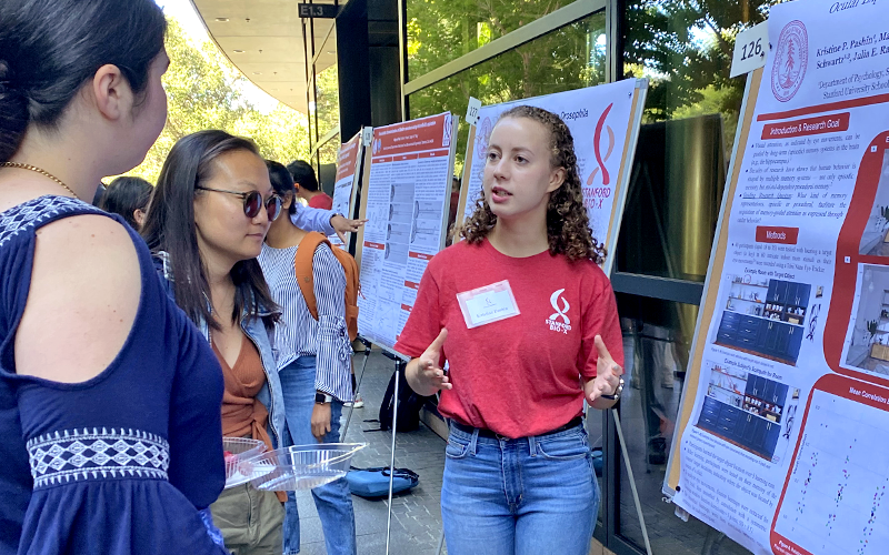 Outdoor photo of a white female undergraduate in a red Bio-X T-shirt showing a poster to other researchers.