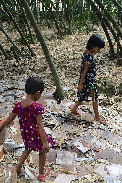 Photo of two young girls wearing brightly-colored dresses walking over a pile of square-shaped scraps of metal.