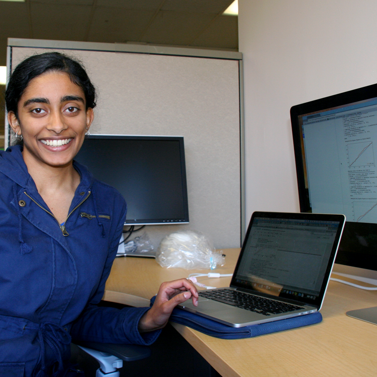 Photo of USRP student Maya Varma in the laboratory, showing graphs on two computer screens.
