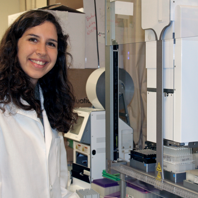 Photo of graduate student Mira Moufarrej in the lab, standing beside a large machine with samples in it.