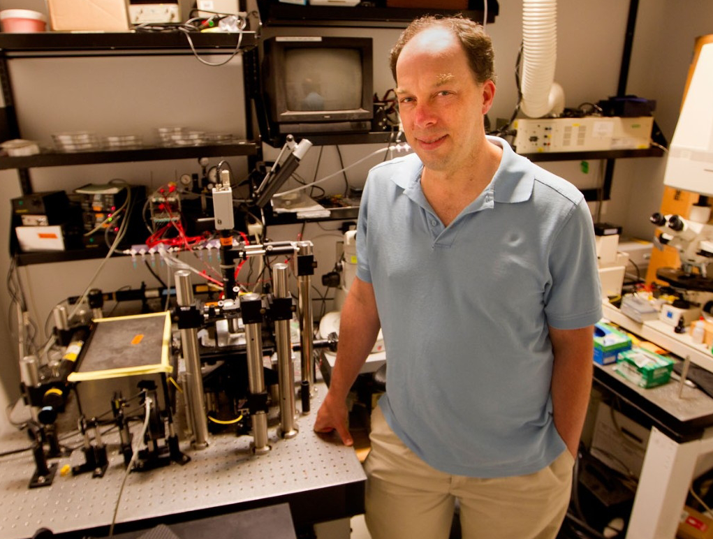 Photo of mid-aged professor in polo shirt standing by advanced lab equipment.