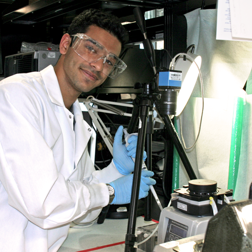 Photo of undergraduate student Rahul Shiv in the lab using a long pipette near several lab equipment devices.