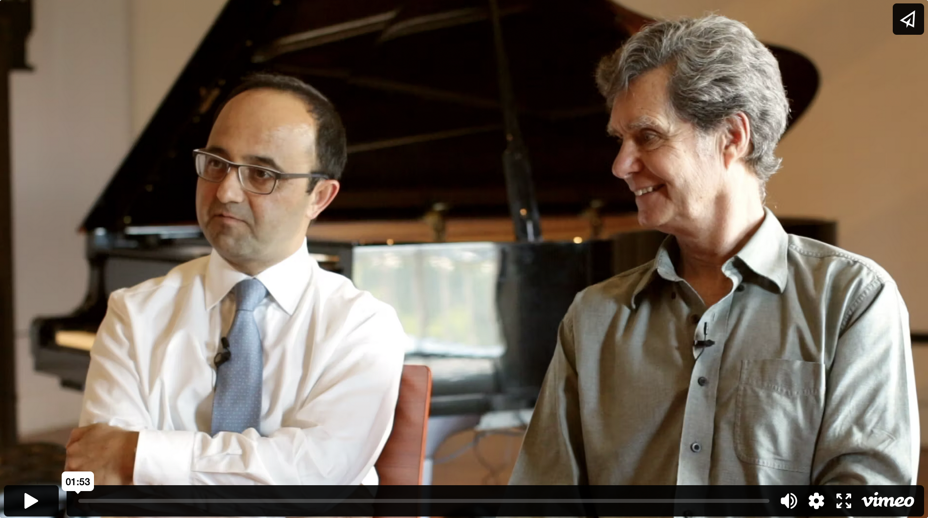 Video player screenshot of two men sitting indoors in front of a piano.