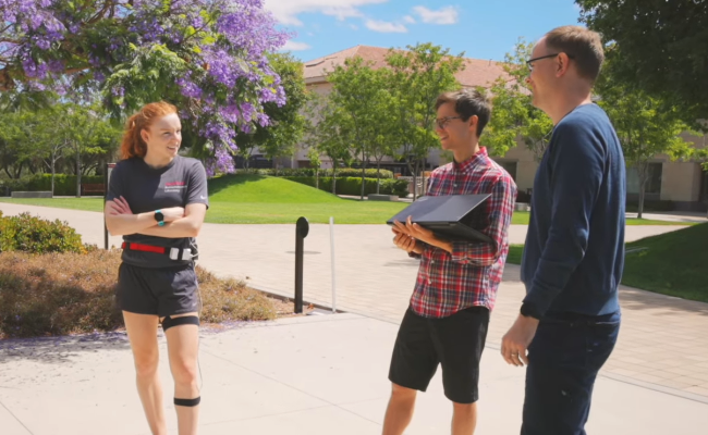 Screenshot of white female graduate student wearing running clothes and the wearable measurement device components, white male graduate student holding a laptop, and white male faculty member standing in an open courtyard area.