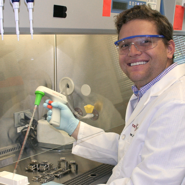Photo of graduate student Aaron Wilk in a wet lab, wearing glasses and a lab coat and gloves, using a long pipetter in a fume hood.