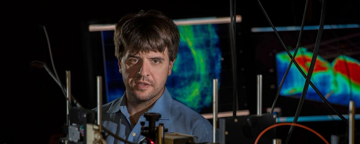 Photo of Dr. Karl Deisseroth in the laboratory, surrounded by equipment with wires and screens displaying brightly-colored optogenetic displays.