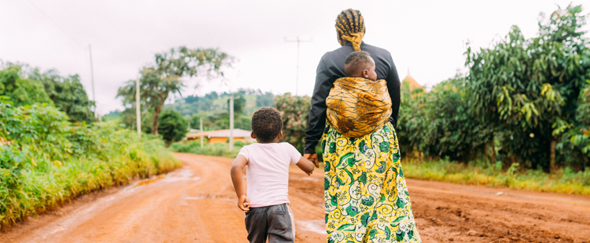 Photo of Kenyan woman walking down a dirt road, with one small child carried in a sling on her back, holding the hand of a slightly older child.