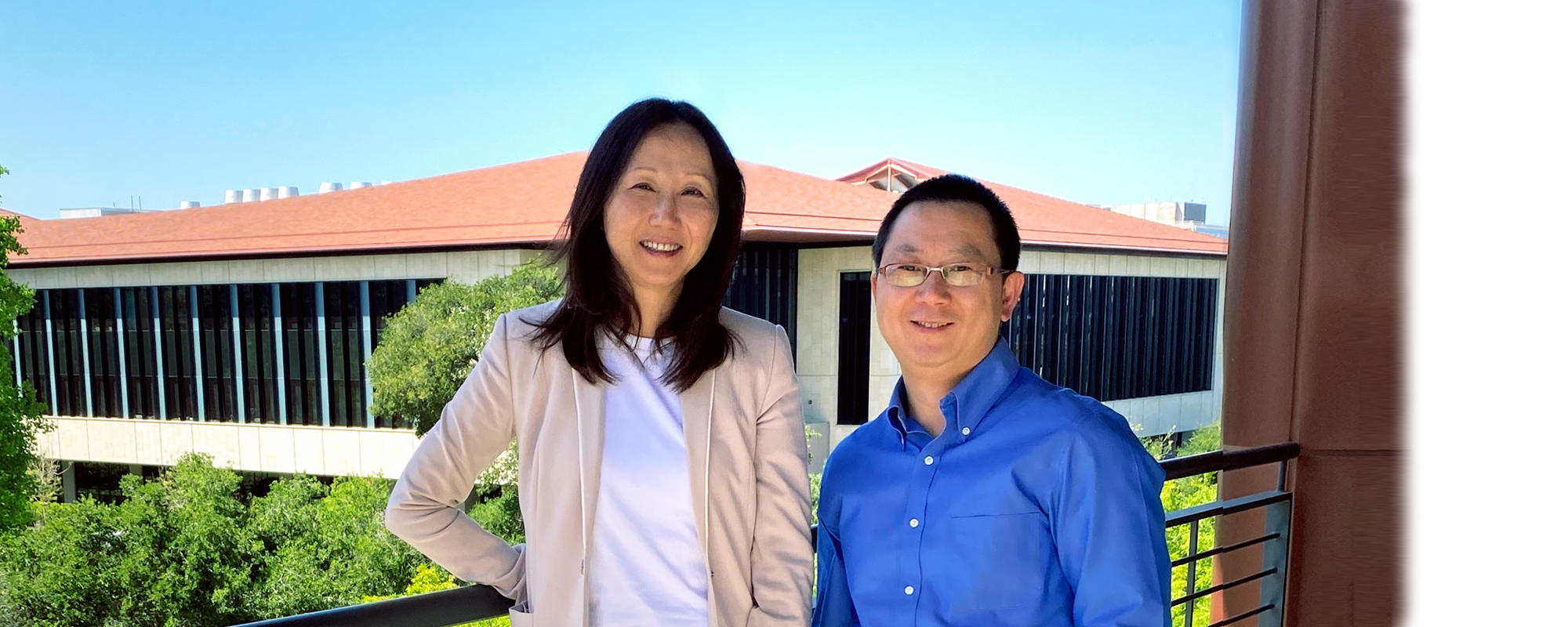 Outdoor photo on a balcony of a smiling Asian female faculty member wearing a pink blazer and a white shirt, with long black hair, and a slightly shorter, smiling male Asian faculty member wearing glasses and a royal blue button up shirt. In the distance behind them are bright green trees and a building with many windows and a red brick roof.