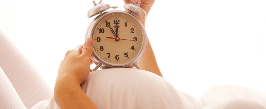 Photo of a pregnant woman lying on her back, holding an old-fashioned alarm clock on top of her stomach.