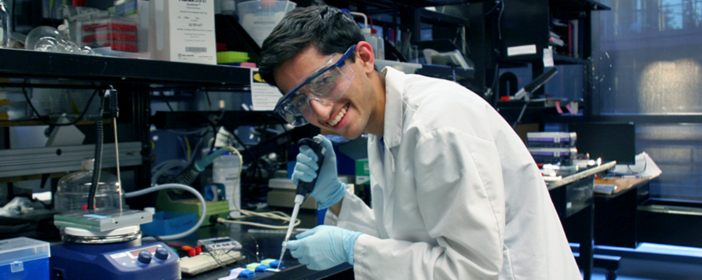 Photo of high school-aged researcher Rohan Mehrhotra in the wet lab space using a micropipette and wearing a lab coat and safety goggles.