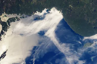 Satellite image of a large body of water and the nearby land, with numerous clouds rippling in different ways.