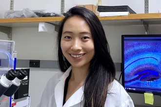 Indoor photo of a smiling Asian female graduate student standing in a wet laboratory next to a microscope, with a computer screen displaying cells in the background.