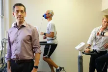 Photo of Dr. Euan Ashley standing at the front of a small room where two subjects, one male and one female, are exercising while wearing multiple kinds of sensors, monitored by a member of the Ashley lab.