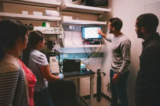 Indoor photo of four diverse lab members gathered around a computer screen, with one male graduate student gesturing to the display.