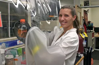 Indoor photo of a white female graduate student with her arms put into a safety hood with plastic, holding up a bottle inside the hood, and smiling at the camera.