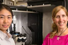 Photo of a young Asian woman and her female Principal Investigator, standing in a specialized lab space with a microscope.