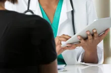 Photo over a female patient's shoulder of a female doctor holding up a clipboard as they talk across a table.
