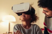 Photo of a young African American girl wearing a VR headset, sitting down with her father.