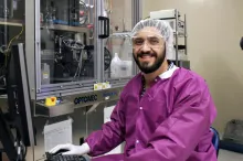 Photo of young male graduate student working in a clean room, gowned and wearing a cap while working on a computer.