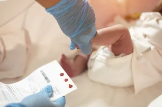 Photo of gloved hand holding a small baby's foot and other hand holding a chart with dots of blood.
