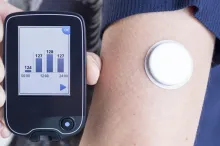 Photo of a woman's arm which bears a glucose monitor, holding up a readout.